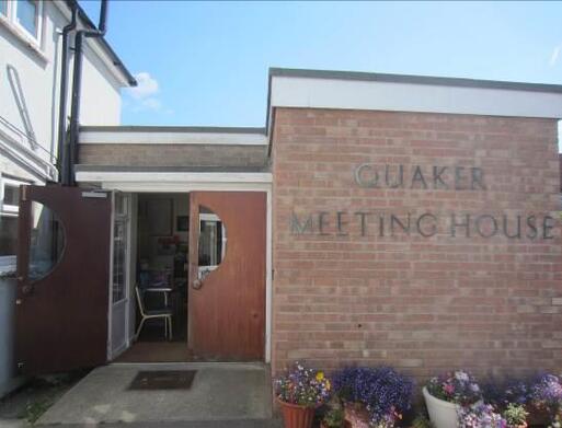 Small brick building with 'Quaker Meeting House' lettering, flowering plants in large pots sit alongside the entrance doors.  