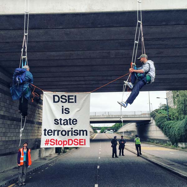 two men abseiling from bridge holding banner says DSEI is state terrorism  #StopDSEI