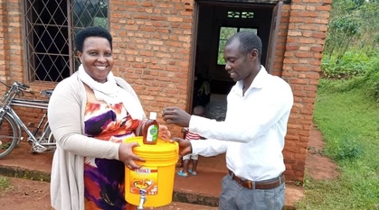a woman holds a bucket with a tap at the bottom and a bottle of dettol sit on top of the sealed bucket. A man is washing his hands.