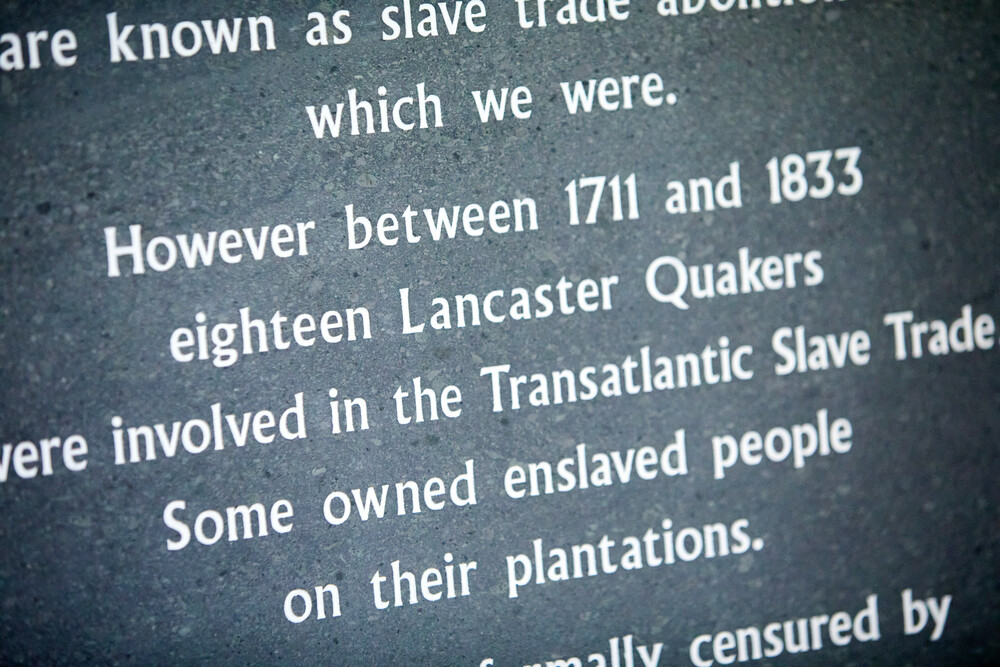 Focus on grey plaque in Lancaster Meeting House. Plaque reads 'Quakers are known as slave trade abolitionists, which we were. However, between 1711 and 1833, 18 Lancaster Quakers were involved in the Transatlantic slave trade. Some owned enslaved people on their plantations. ...'