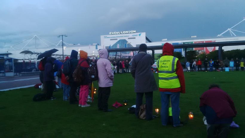 A circle of people of many faiths and none stand in a circle by candlelight outside the DSEI Arms Fair at London's Excel Centre.