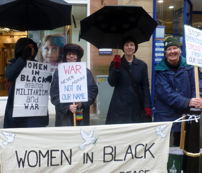Four women in black and navy standing in the rain with black umbrellas holding a vigil for peace