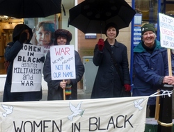 Four women in black and navy standing in the rain with black umbrellas holding a vigil for peace