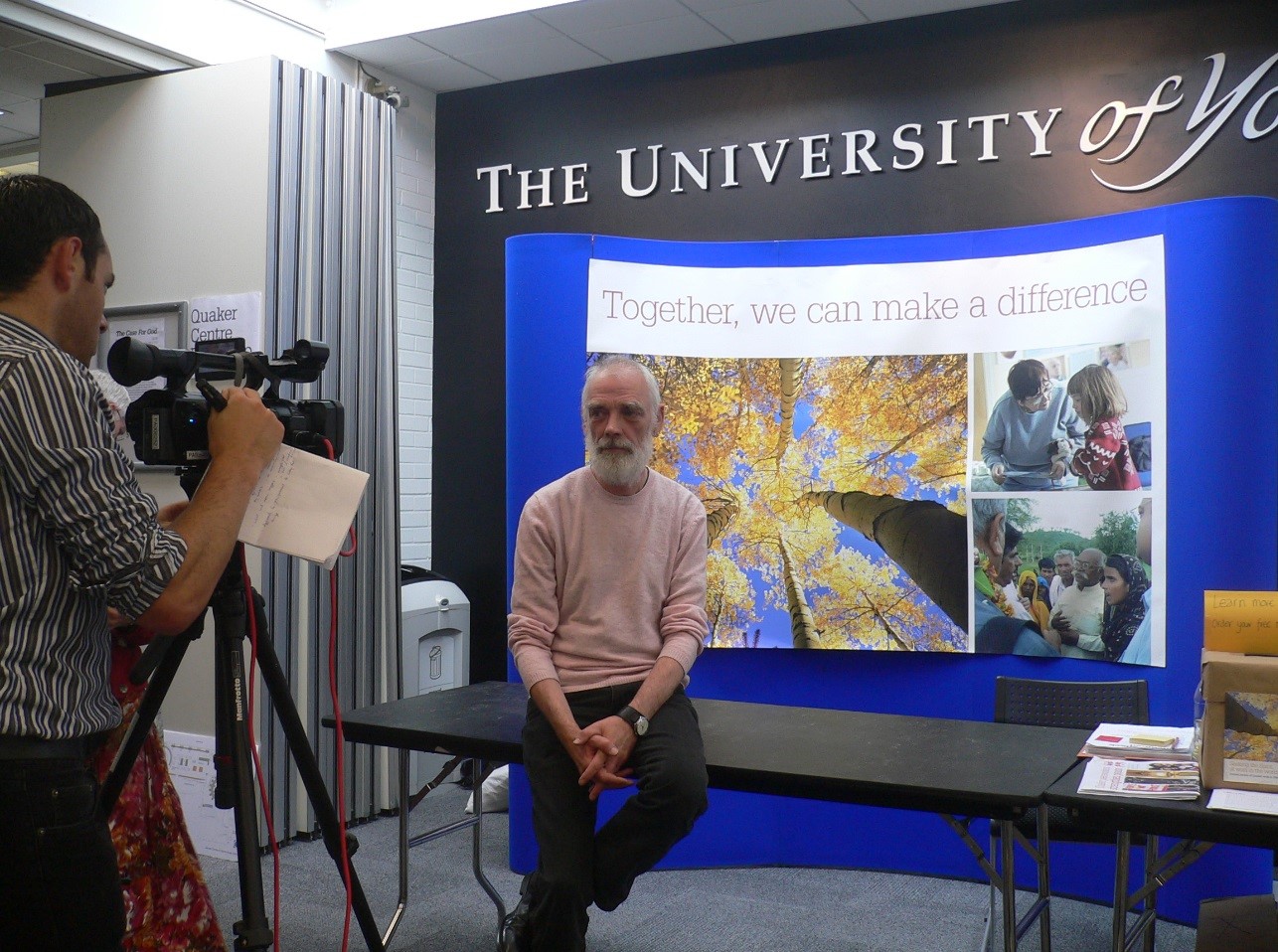 Seated man being filmed in front of Quaker poster