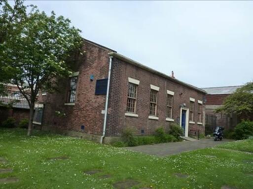 Tall brick building with five long sash windows and a slate roof. The entrance is accessed by a garden path. 