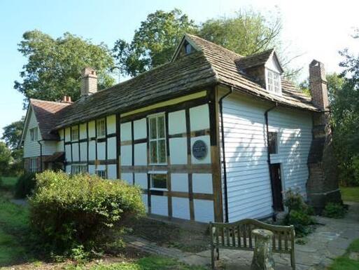 Sixteenth century white and timber-framed farmhouse set within garden grounds. A blue plaque is affixed to the right side of the building.