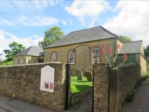 Short stone building and burial ground viewed through a metal archway set within high stone boundary walls. 