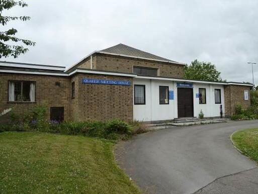 Wide brick building with circular road to large white-painted entrance, blue 'Quaker Meeting House' sign affixed to front wall. 