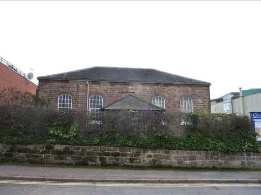 Stone building with large windows is partially concealed from the street by a large hedge and a stone wall.