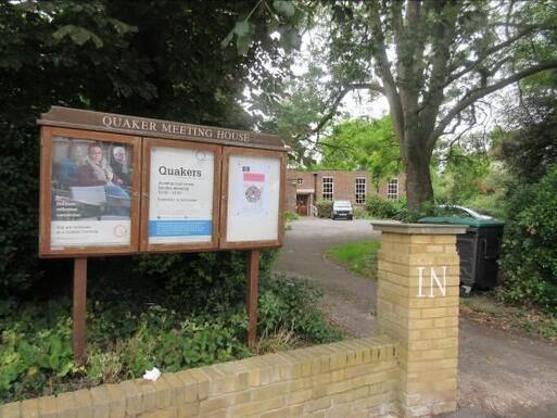 Short brick building with large windows and car park, a large wooden Quaker Meeting House noticeboard is at the front of the grounds.