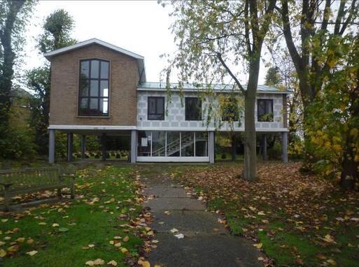 Brick building raised on plinths above a large garden, steps lead up into the entrance. 