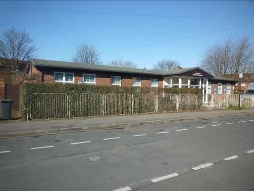 Single storey building with wide windows is hidden from the road by a large boundary hedge. 