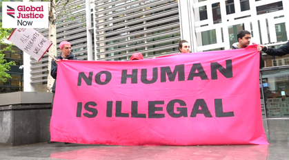 5 people holding up a bright pink banner outside on a grey day with 'No Human Is Illegal' on it. 