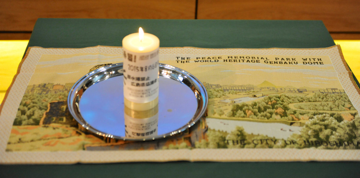 A lit candle on a silver plate sitting on top of a tapestry of the Hiroshima memorial peace garden