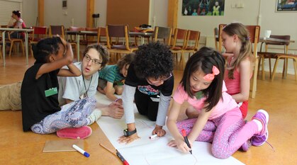 five children on the ground drawing on a large sheet of paper with felt tips. An adult is laying on the ground talking to one of them.