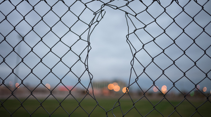 a small hole in chain link fence leading to an unclear and murky field beyond