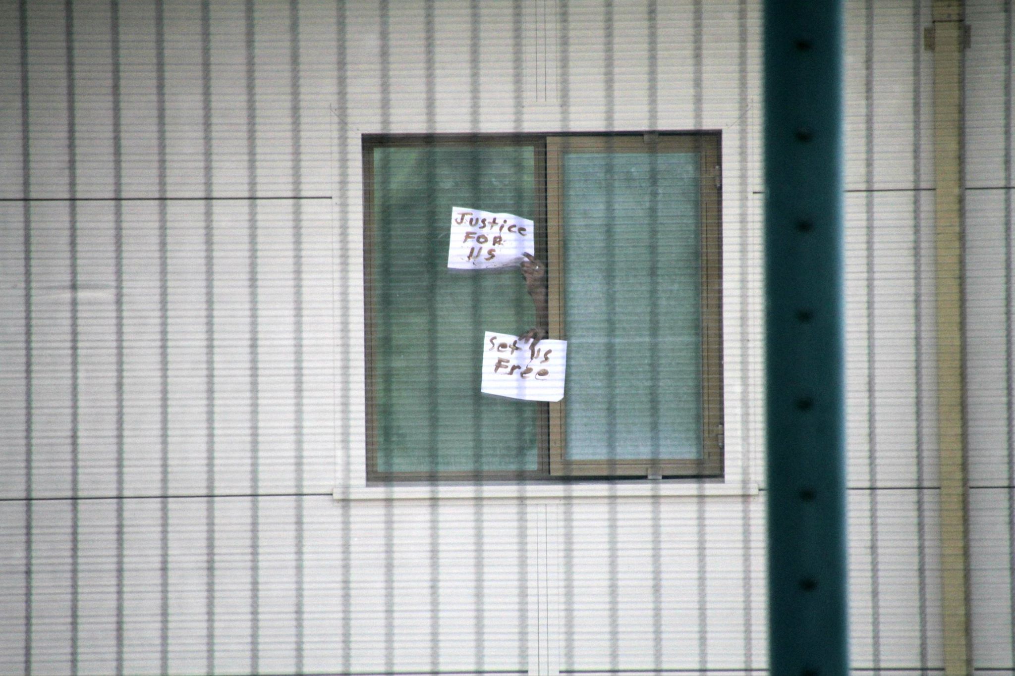 two hands coming out of a window of an immigration detention centre holding up pieces of paper with the words 'justice for us' 'set us free' written on it