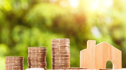 three piles of coins and wooden model of a house on a sunny window ledge