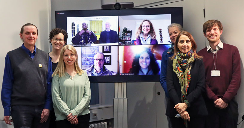 People standing on either side of a screen with Zoom faces on it 