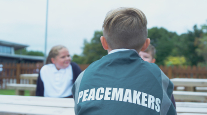 three children sat around a bench outside. One is wearing a tabard that says peacemaker