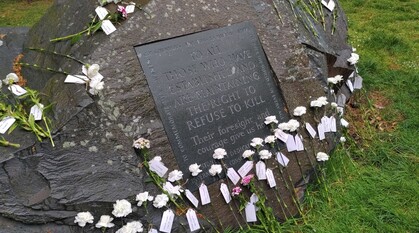 Wet conscientious objectors' memorial stone covered by white carnations. Plaque reads: to all those who have established and are maintaining the right to refuse to kill. Their foresight and courage give us hope.