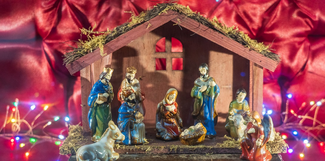 A nativity scene with a wooden stable and china figures set against a background of a silky red material with fairy lights at the corners.