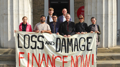 A group of people on some church steps holding a banner saying 'loss and damage finance now'