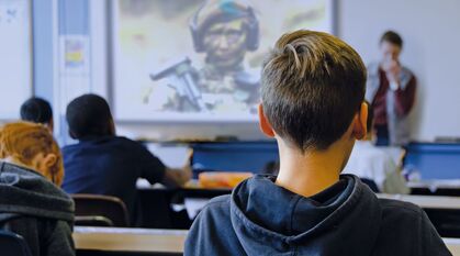 Children sat at desks in a classroom watching a video of a soldier in camo on a projector screen