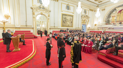 Leasa Lambert standing in nice but ordinary clothes in an opulent room with red carpet and gilded cornices ready to address Charles Windsor. Others in the room are wearing more ceremonial robes – from full academic dress to clerical garb