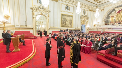 Leasa Lambert standing in nice but ordinary clothes in an opulent room with red carpet and gilded cornices ready to address Charles Windsor. Others in the room are wearing more ceremonial robes – from full academic dress to clerical garb