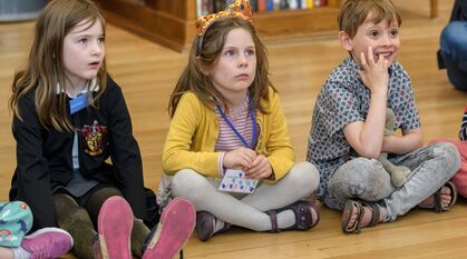 Three children sitting cross legged on a wooden floor enthralled by something out of shot