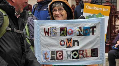 A person in a rain coat at a protest holds up a handstitched banner quoting a section of Quaker Advices & Queries: 'We do not own the world...'