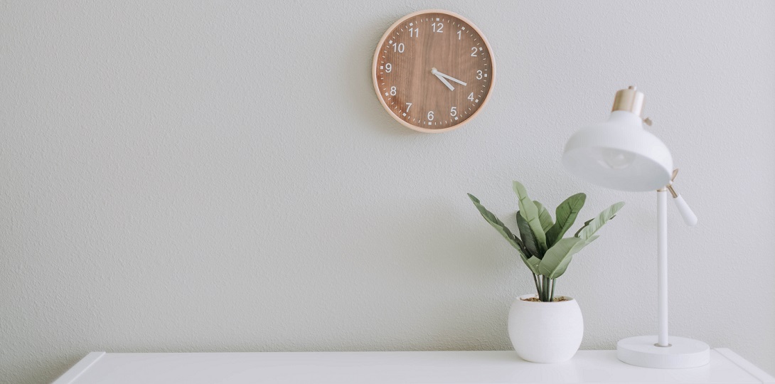 A photograph of a plain white desk with a plant pot, a lamo, and a clock