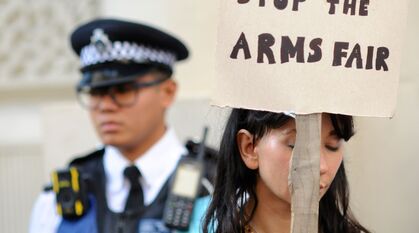A Quaker protestor with a placard that says 'stop the arms fair'. Behind her is a police officer.