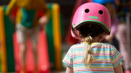 child in a pink helmet cycling along a pavement, in the distance another child is playing on a slide