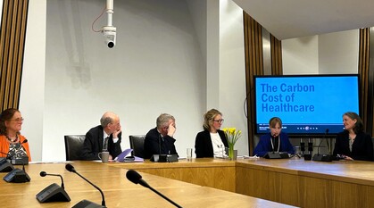 A panel of speakers in a room in the Scottish Parliament. A screen behind the speakers says 'The Carbon Cost of Healthcare'