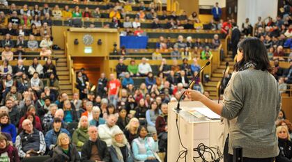 A person speaking from a lectern looking out over an audience in raised seating in The Light in Friends House