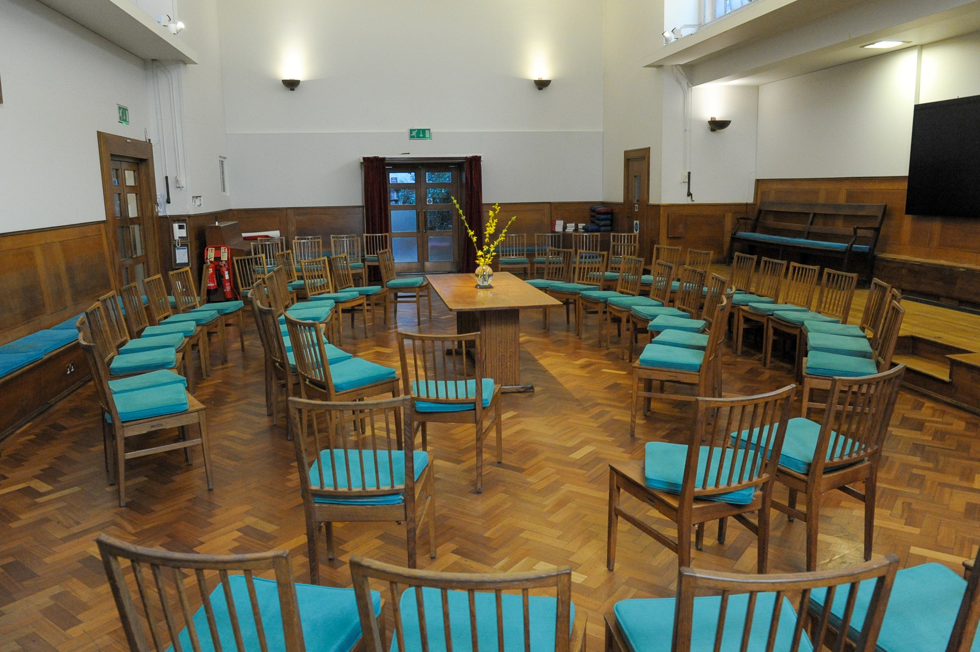 The main room used by Westminster Quakers for meetings for worship. Light comes in from the windows above to the wooden chairs with their teal blue seat covers arranged in a circle. A vase of tall yellow flowers sits in the centre of the room