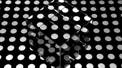 A clear glass cube sits on a black background with white lit polka dots, such that the pattern is reflected, repeated, and broken up by the cube - otherwise, it would be invisible.