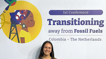 Daniela Campos standing in front of a sign with the logo of the conference (a colourful bird in flight and a woman walking peacefully away from drilling equipment) and the title saying '1st conference: Transitional away from fossil fuels – Colombia & The Netherlands