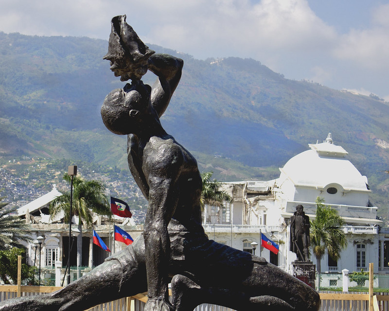 A monumental statue of a freedman stands triumphantly in the foreground, raising a conch shell to his lips and sounding it in celebration of freedom and resilience. Behind him is a stately white building , framed by Haitian flags. In the distance the hills of Haiti stretch across the horizon.