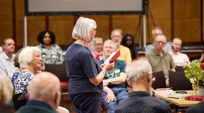 A person stands to read from Advices & Queries in a Quaker meeting for worship. Worshippers are sat in a circle around her listening, some with eyes closed. Other worshippers are visible online.